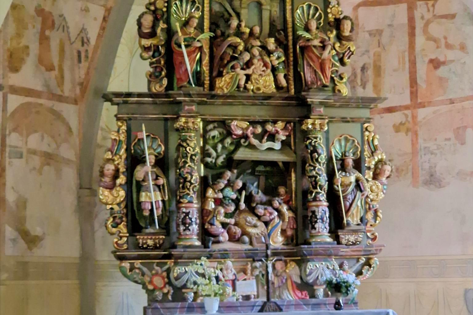 Stellen Sie sich vor, Sie stehen oder sitzen in einer kleinen Kapelle. Vor Ihnen befindet sich ein reich verzierter, farbig gefasster Altar aus Holz. Der Altar ist mehrstöckig aufgebaut und wirkt wie ein kleines, kunstvolles Haus mit Säulen, Figuren und Nischen. Die Farben sind warm: Gold, Rot, Grün und dunkles Holz prägen das Gesamtbild. Im Zentrum des Altars, auf Augenhöhe, sehen Sie eine Darstellung der Geburt Jesu: Ein kleines Kind liegt in der Mitte, umgeben von mehreren Figuren. Einige Figuren knien, andere stehen. Alle sind dicht um die Mitte gruppiert, sodass ein Gefühl von Nähe und Sammlung entsteht. Darüber, auf einer erhöhten Ebene, befinden sich weitere Figuren, die Szenen aus dem Leben Jesu oder aus der biblischen Geschichte zeigen. Diese Figuren sind kleiner, aber ebenfalls reich gestaltet. Auch hier stehen mehrere Gestalten beieinander, eingerahmt von Säulen und Bögen. Seitlich stehen größere Figuren in Nischen. Jede Figur ist einzeln hervorgehoben, mit Gewändern in kräftigen Farben und goldenen Details. Unterhalb der Hauptszene gibt es eine weitere kleine Darstellung, ebenfalls mit mehreren Figuren, die sich um eine zentrale Szene gruppieren. Vor dem Altar steht ein schlichter, heller Altartisch mit einem weißen Tuch. Darauf befinden sich frische Blumen. Im Hintergrund erkennt man die Wände der Kapelle mit zarten, teilweise verblassten Malereien. Die Decke ist gewölbt und trägt ebenfalls helle, zurückhaltende Bildmotive. Insgesamt entsteht ein Eindruck von Stille, Konzentration und Tiefe: Der kunstreiche Altar lädt dazu ein, den Blick immer wieder zur Mitte zu führen – dorthin, wo das Wesentliche liegt.