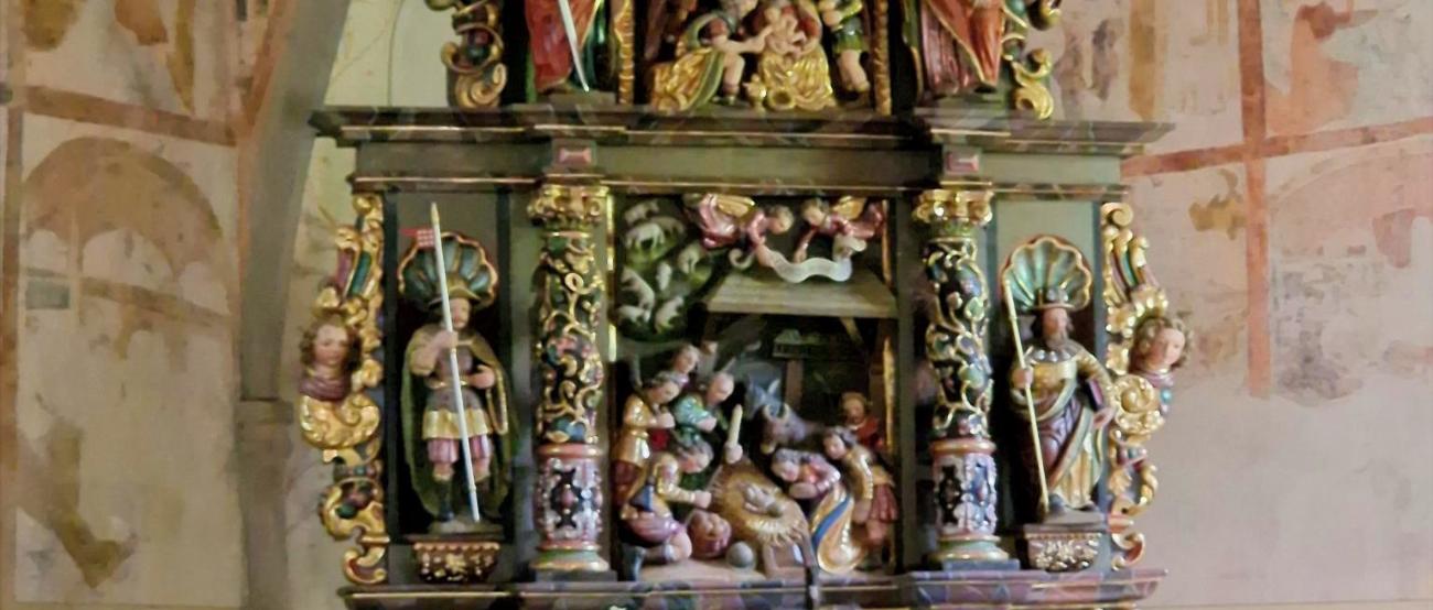 Sankt Jost_Altar_Andachten (c) Hans Georg Bach Stellen Sie sich vor, Sie stehen oder sitzen in einer kleinen Kapelle. Vor Ihnen befindet sich ein reich verzierter, farbig gefasster Altar aus Holz. Der Altar ist mehrstöckig aufgebaut und wirkt wie ein kleines, kunstvolles Haus mit Säulen, Figuren und Nischen. Die Farben sind warm: Gold, Rot, Grün und dunkles Holz prägen das Gesamtbild. Im Zentrum des Altars, auf Augenhöhe, sehen Sie eine Darstellung der Geburt Jesu: Ein kleines Kind liegt in der Mitte, umgeben von mehreren Figuren. Einige Figuren knien, andere stehen. Alle sind dicht um die Mitte gruppiert, sodass ein Gefühl von Nähe und Sammlung entsteht. Darüber, auf einer erhöhten Ebene, befinden sich weitere Figuren, die Szenen aus dem Leben Jesu oder aus der biblischen Geschichte zeigen. Diese Figuren sind kleiner, aber ebenfalls reich gestaltet. Auch hier stehen mehrere Gestalten beieinander, eingerahmt von Säulen und Bögen. Seitlich stehen größere Figuren in Nischen. Jede Figur ist einzeln hervorgehoben, mit Gewändern in kräftigen Farben und goldenen Details. Unterhalb der Hauptszene gibt es eine weitere kleine Darstellung, ebenfalls mit mehreren Figuren, die sich um eine zentrale Szene gruppieren. Vor dem Altar steht ein schlichter, heller Altartisch mit einem weißen Tuch. Darauf befinden sich frische Blumen. Im Hintergrund erkennt man die Wände der Kapelle mit zarten, teilweise verblassten Malereien. Die Decke ist gewölbt und trägt ebenfalls helle, zurückhaltende Bildmotive. Insgesamt entsteht ein Eindruck von Stille, Konzentration und Tiefe: Der kunstreiche Altar lädt dazu ein, den Blick immer wieder zur Mitte zu führen – dorthin, wo das Wesentliche liegt.