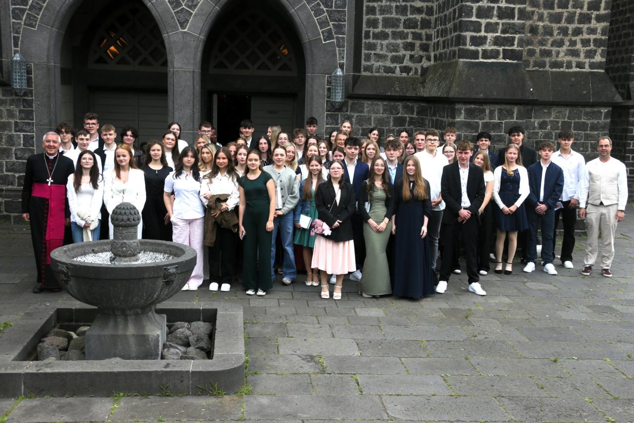 Gruppenfoto mit vielen jungen Menschen vor einer Kirche mit dunklen Steinen.