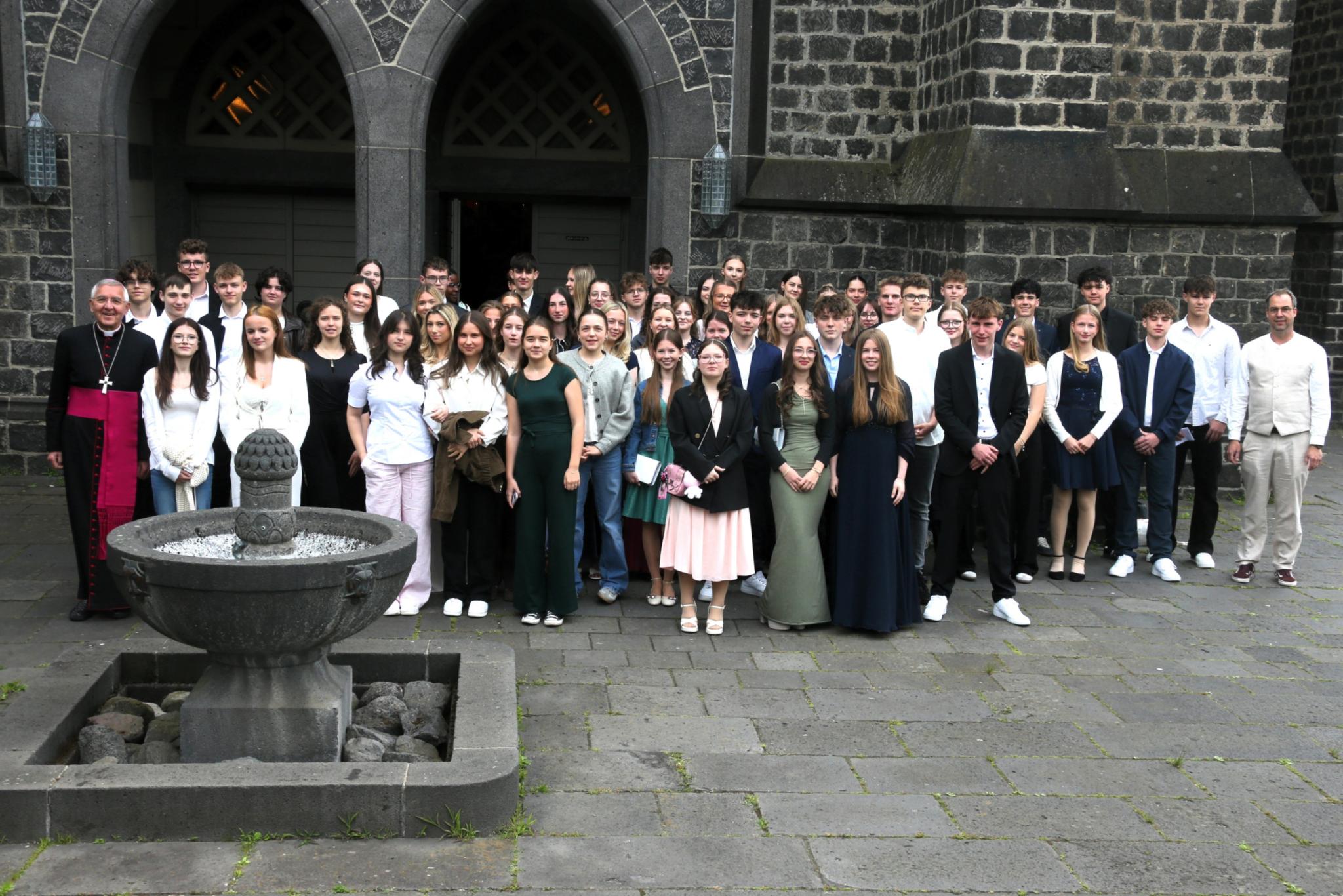 Gruppenfoto mit vielen jungen Menschen vor einer Kirche mit dunklen Steinen.