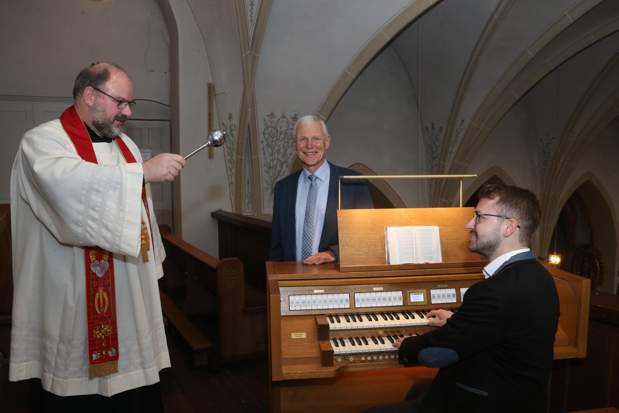 Ein Pfarrer besprengt eine Orgel mit Weihwasser, ein Mann sitzt an der Orgel, ein weiterer Mann steht hinter der Orgel und lächelt.